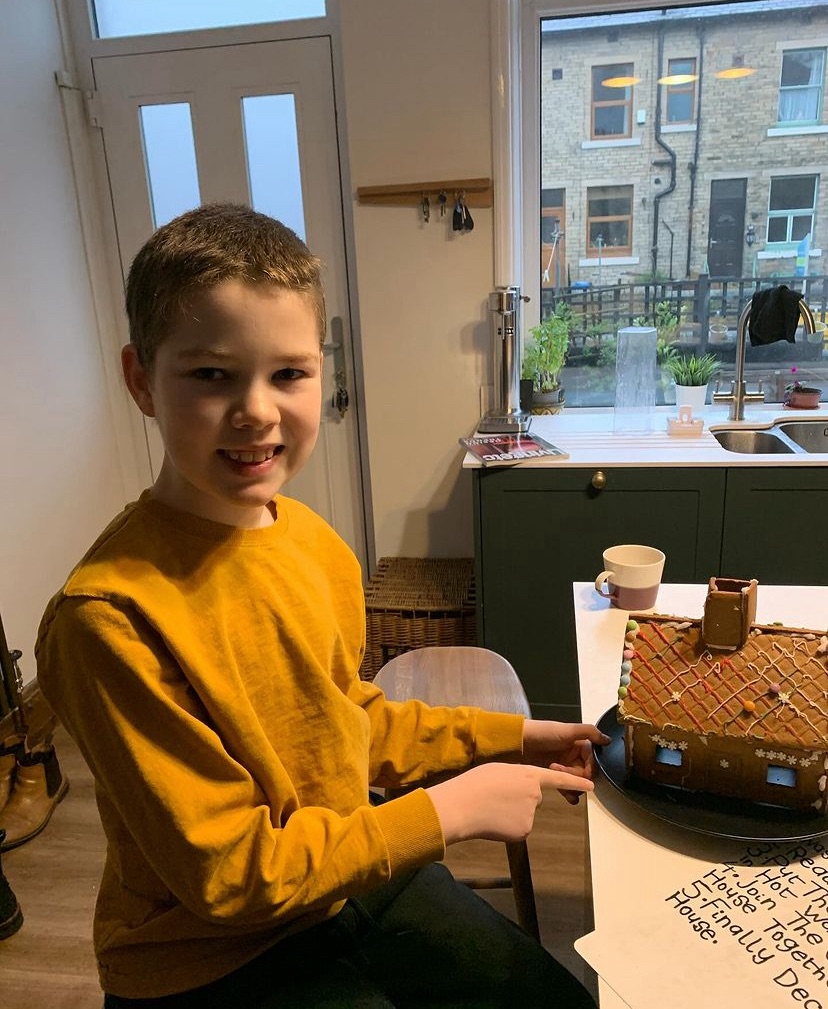 Jake is smiling and pointing to a gingerbread house that he has made. He looks very pleased with himself. 